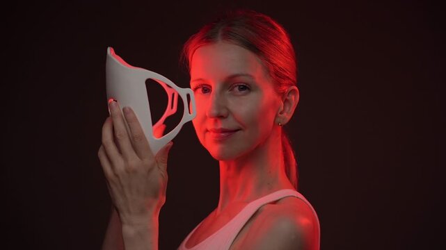 woman holding infrared LED mask profile side view, then turn face to the camera and smiles. biohacker theme professional skin care video footage. black background