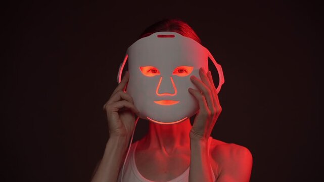 beautiful woman looks at the camera and smiles holding infrared LED mask, then turns in profile to an infrared cosmetic mask. biohacker theme professional skin care video footage. black background