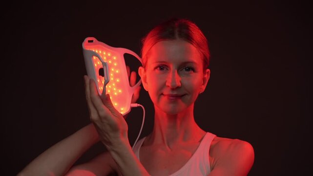 woman holding infrared light therapy LED mask then move it away and shows the face smiling.  relaxed smiling face looking at the camera, black  background. Contrast red light. horizontal composition v