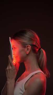 woman using infrared light face mask. infrared light mask.  black background. professional skin care. vertical video footage. relaxed smiling blonde woman