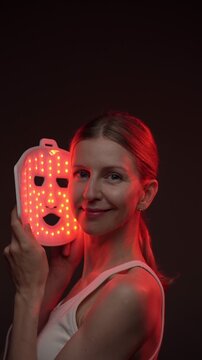 relaxed smiling blonde woman using infrared light face mask. infrared light mask.  black background. professional skin care. vertical video footage.