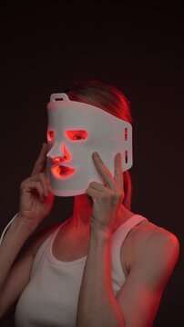 blonde woman using infrared light face mask. infrared light mask.  black background. professional skin care. vertical video footage.