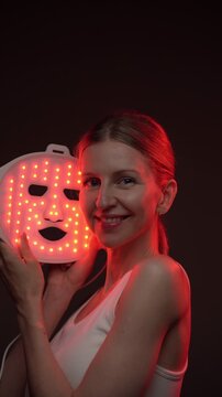woman 37-year-old smiles while using an infrared facial mask. Professional cosmetic skin care. vertical video footage black background, red light