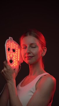 woman 35-year-old smiles while using an infrared facial mask. Professional cosmetic equipment. vertical video footage black background, red light