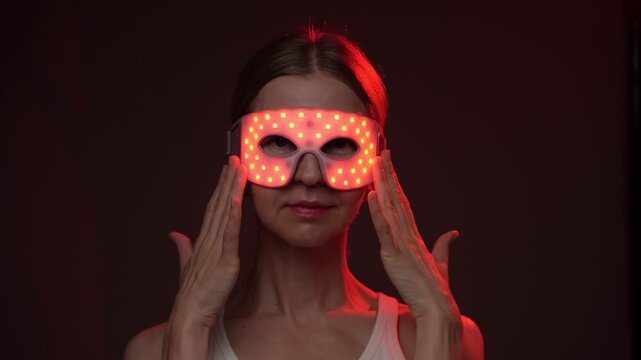 Red and infrared light therapy eye mask. smiling woman wearing infrared light eyes therapy mask. touching the mask with her hand.  looking side. dark background. video footage