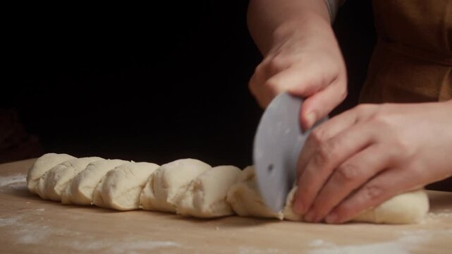 Close-up of hands using a pizza cutter to slice freshly rolled dough into equal portions on a floured wooden board. Traditional bread preparation in a cozy home kitchen.