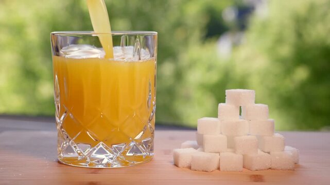 Orange juice is poured into a glass along with a stack of sugar cubes, refreshing beverage. Fructose or sugar, consumed in excess, are unhealthy. Increased risk of diabetes, weight gain, fatty liver.