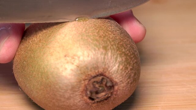 A hand is holding a kiwi on a cutting board and cutting it in half with a knife. It contains vitamin C, fiber, and potassium. Nutrition, fruit, healthy lifestyle, fresh, kitchen.