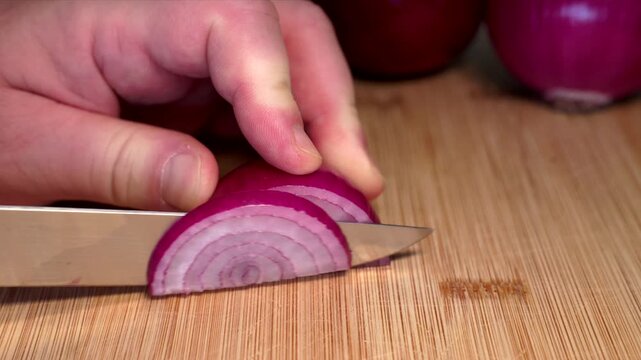 A hand is holding a red onion on a cutting board and slicing it with a knife. Contains vitamin C and fiber. Nutrition, vegetables, healthy lifestyle, fresh, raw food. 