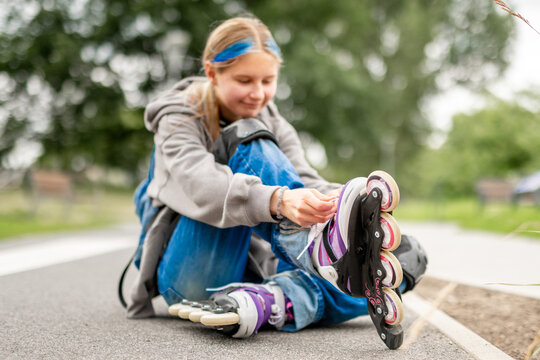 Young girl sits on paved ground, adjusting her purple and white rollerblade during daytime.