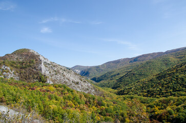 Fototapeta premium Scenic mountain valley with rocky slopes, forested hills and vibrant autumn colors beneath a clear blue sky. 
