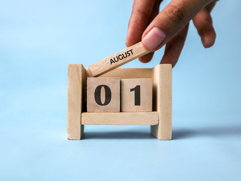 A woman's hand holds a wooden block with August written on it on a wooden block calendar for August 1st against a light blue background.