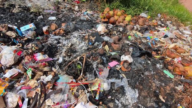 Video of Ash and charred waste lies scattered. Broken plastic and burnt packaging mix with coconut husks. Burned items rest near unburned coconuts and green grass. 4K Footage at a rubbish dump site