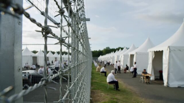 Barbed wire fence bordering a refugee camp with white tents and people sitting outdoors, eye level