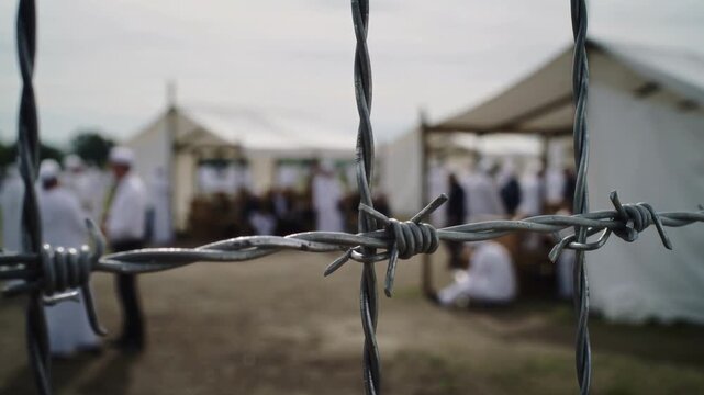 Barbed wire fence with blurred refugee camp tents in background, extreme close up shot