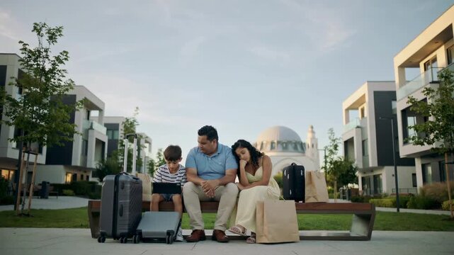 Middle Eastern family sitting on bench with suitcases and tablet outdoors with copy space eye level