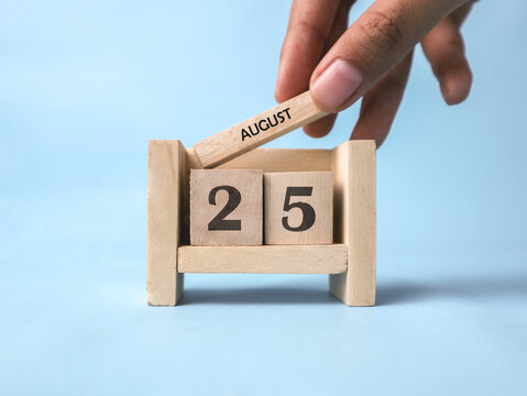 A woman's hand holds a wooden block with August written on it on a wooden block calendar for August 25th against a light blue background.