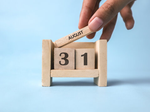 A woman's hand holds a wooden block with August written on it on a wooden block calendar for August 31st against a light blue background.