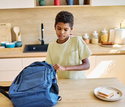 Portret of  a black teen boy with a school backpack having fun alone preparing sanwiches snack for  breakfast and school  in kitchen