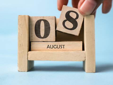 A woman's hand is holding a wooden block with the number 8 written on it on a wooden block calendar for August 8th on a light blue background.