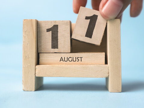 A woman's hand is holding a wooden block with the number 11 written on it on a wooden block calendar for August 11th on a light blue background.