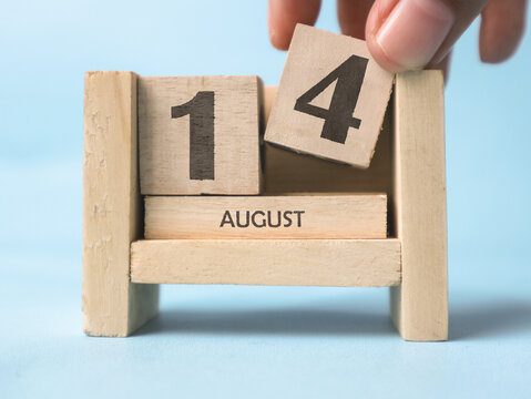 A woman's hand is holding a wooden block with the number 4 written on it on a wooden block calendar for August 14th on a light blue background.