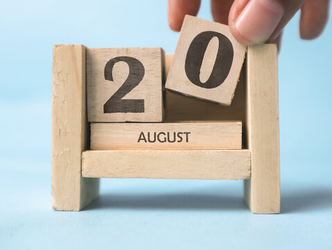 A woman's hand is holding a wooden block with the number 0 written on it on a wooden block calendar for August 20th on a light blue background.