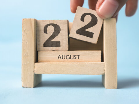 A woman's hand is holding a wooden block with the number 2 written on it on a wooden block calendar for August 22nd on a light blue background.