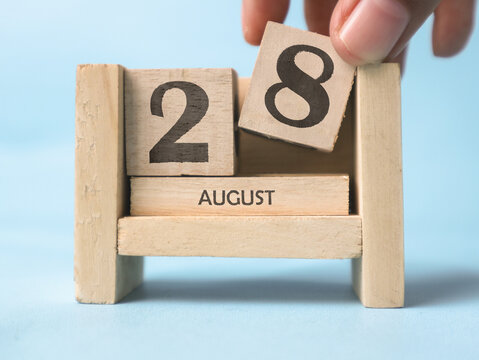 A woman's hand is holding a wooden block with the number 8 written on it on a wooden block calendar for August 28th on a light blue background.