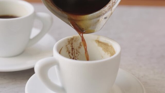 Hot, strong traditional turkish coffee being poured from a cezve into a small white ceramic cup