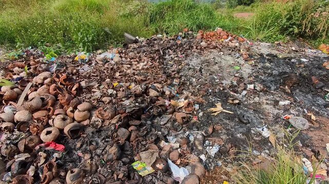 Video of Discarded coconut shells and debris litter a muddy roadside patch. Green grass grows nearby, although thick trash dominates the ground. Burnt earth and broken ceramics hint at recent fire or 