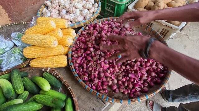 Video of a traditional market vendor stirring a pile of shallots. Hand-selecting pink shallots from a large woven basket. Fresh cucumbers, corn, and garlic complete the colorful display. 4K footage 