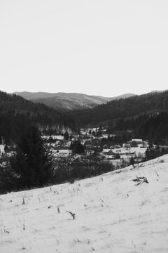 Winter landscape shows village in snowy valley beneath dark forests and distant hills under pale sky glow