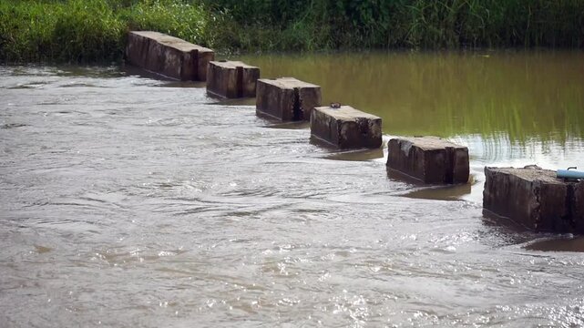 Muddy river water flowing rapidly through a small concrete dam barrier. Rural irrigation system and water management infrastructure footage showcasing stream current movement in countryside