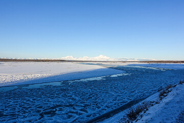Ice pans drift down a broad braided Alaskan river across snowy gravel bars, with the white peaks of Denali and the Alaska Range on the far horizon under a deep blue sky. © Benedikt