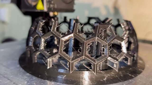 A modern 3D printer nozzle precisely deposits black plastic filament to construct a complex geometric hexagonal mesh object on the build plate.