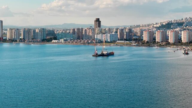 Dredging barge and support vessel in calm bay with buildings on shoreline. Aerial footage of seabed cleanup and coastal maintenance activity