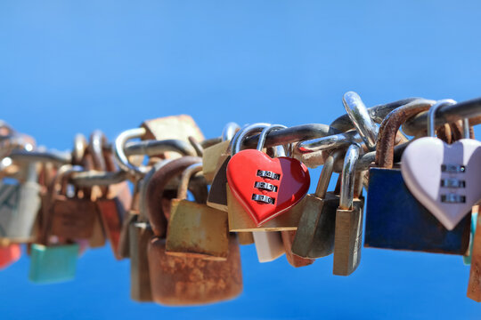 Red heart shaped combination love lock attached to chain with other padlocks against blue sky. Concept for everlasting love, romance, Valentine day, commitment, and relationship.