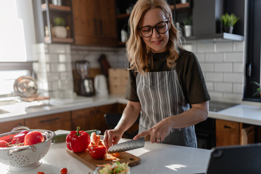 Woman preparing vegetables while attending a video call on a tablet in a bright kitchen. Concept of remote communication, online consultation, multitasking and modern lifestyle