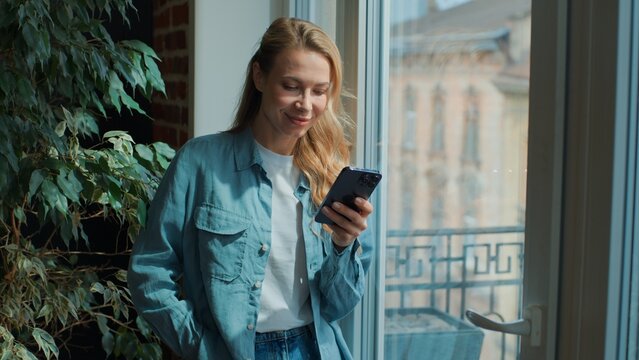 Happy Caucasian middle-aged woman using mobile phone looking at window smiling inspired chatting with smartphone at home adult female browsing social media in cellphone app look outside in apartment