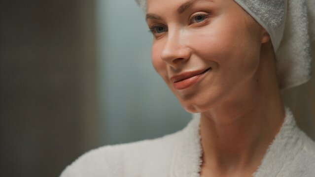 Close up Caucasian middle-aged woman wraped hair in towel looking at mirror smiling happy pampering in bathrobe adult female enjoying hairstyle procedure skincare routine smile cheerful in morning