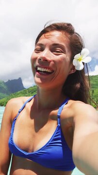 Selfie. Tropical vacation bikini woman smiling at camera at luxury travel resort. Happy girl relaxing with polynesian flower, Asian beauty. Raw vertical video of real woman without any makeup