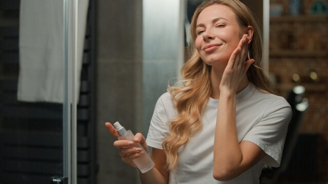 Smiling carefree Caucasian middle-aged woman applying spray thermal water lotion serum toner for face refreshing facial skin happy female smile spraying micellar product beauty routine at bathroom