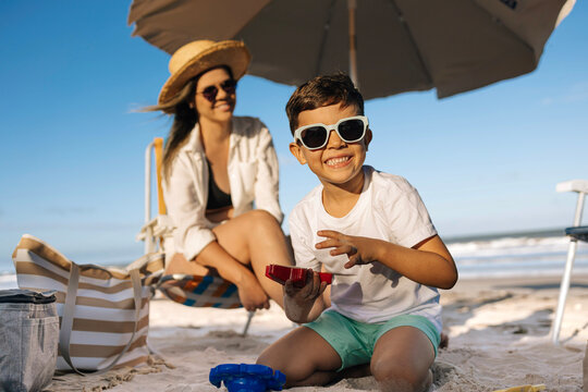 Mother and Son Playing on Sandy Beach Under Umbrella During Sunny Summer Vacation Together