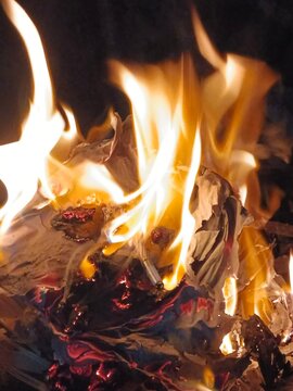 Api menyala terang membakar kayu dan kertas hingga menghasilkan bara merah yang menyala. Lidah api berwarna kuning dan oranye terlihat bergerak dinamis, menciptakan suasana hangat dan dramatis. Foto d