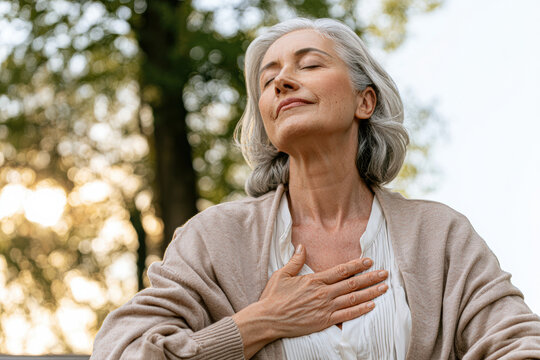 Mature woman with closed eyes, hand on chest, appreciating nature in soft sunlight among blurred leafy trees.