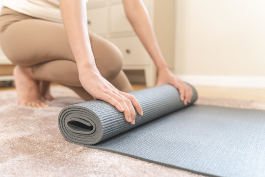 Close up of an Asian young woman, girl rolling up a grey mat after a yoga or fitness workout session on a carpet at home, healthy lifestyle routine and mindfulness self-care concept.
