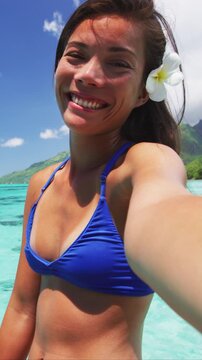 Bikini woman blowing a kiss smiling at camera at luxury travel resort. Happy girl relaxing with polynesian flower, Asian beauty. Raw vertical video of real woman without any makeup