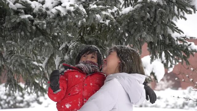 Happy Asian mother holding her son in red jacket under snow-covered pine tree.