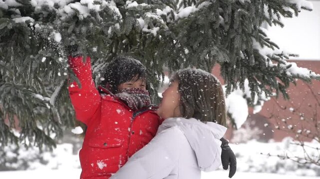Happy Asian mother holding her son in red jacket under snow-covered pine tree.